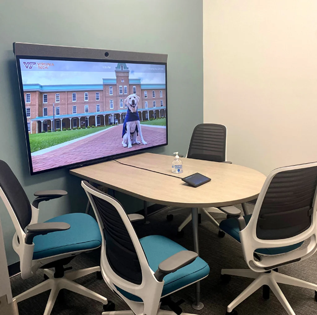A camera on LCD screen on wall with four chairs and a table in huddle room.